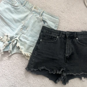 Två pack gögmidjade jeansshorts med fransar - Säljer två par snygga jeansshorts, ett par ljusblå och ett par svarta. Båda har råa, fransiga kanter och klassisk femficksdesign. Perfekta för sommardagar och festivaler. Passar dig som gillar en avslappnad och trendig stil.