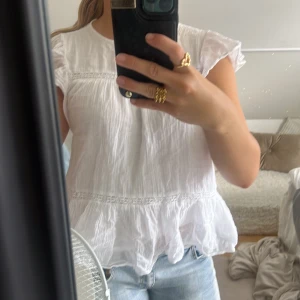 Vit blus med volangdetalj - Supersöt vit blus med volang nertill och fina spetsdetaljer. Blusen har korta ärmar och en luftig, lätt struktur som passar perfekt till jeans eller kjol. Enkel och fräsch stil som känns både romantisk och trendig.