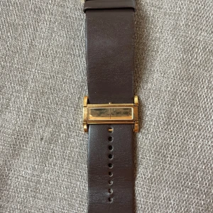 Guldig CK klocka med bredt läderband - Snygg och unik klocka från Calvin Klein med rektangulärt guldigt ur och bredt mörkbrunt läderarmband. Minimalistisk design med smal urtavla och diskreta visare. Perfekt för dig som gillar stilrena accessoarer med en twist.