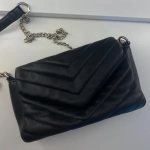 Svart quiltad axelväska med kedja - Snygg stockholmstil svart axelväska i quiltad design med silvrig kedjerem. Väskan har ett stilrent mönster i v-format och är gjord i mjukt skinnliknande material. Den är i superbra skick och passar perfekt till vardagliga outfits! 