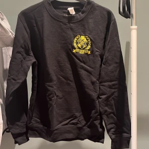 Svart AIK crewneck med broderad logga - Svart crewneck-tröja från AIK med broderad gul AIK Norra Stå-logga på bröstet och schackrutigt märke i nacken. Klassisk passform och rund halsringning. Perfekt för dig som vill visa ditt AIK-stöd på ett stilrent sätt.
