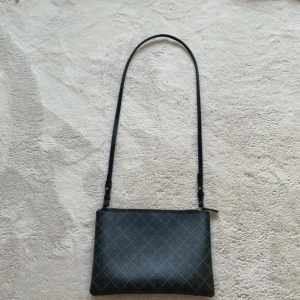 Svart axelväska med rutigt mönster - Stilren svart axelväska med långt justerbart axelband och diskret rutigt mönster. Väskan har guldfärgad dragkedja och är tillverkad i ett tåligt skinnliknande material. Perfekt storlek för det viktigaste och enkel att matcha till olika outfits.