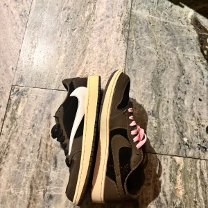 Nike Air Jordan 1 Low Travis Scott - Nike Air Jordan 1 Low Travis Scott med bruna och svarta paneler i skinn och mocka, vit omvänd swoosh och kontrasterande rosa och svarta skosnören. Röd broderad logga på hälen och beige sula ger en unik look. Extra skosnören medföljer. Storlek 44 men lite mindre i storleken