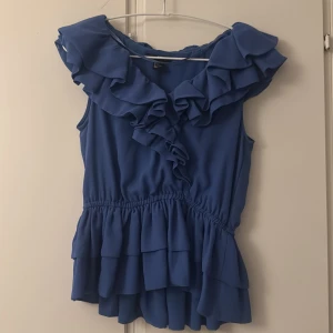 Blå volangblus med ärmlös design - Supersöt blå blus med volanger längs ringningen och nertill. Ärmlös modell med markerad midja och flera lager volanger som ger en cool  look. Perfekt för dig som vill sticka ut lite extra med en gullig topp.