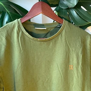 Olivgrön t-shirt med broderad detalj - Olivgrön t-shirt i mjuk bomull med rund halsringning och korta ärmar. På bröstet finns en liten broderad detalj med romerska siffran II i gult. Enkel och clean design som passar till allt.