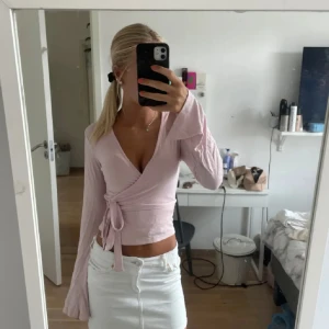 Ljusrosa omlottopp med knyt - Supersöt ljusrosa omlottopp med långa ärmar och v-ringning. Toppen har knyt i sidan och är figurnära med lite utsvängda ärmslut. Perfekt för dig som gillar en feminin och trendig stil.