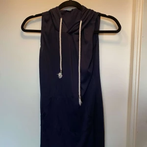 Marinblå ärmlös hoodieklänning - Snygg marinblå ärmlös klänning med huva och vita snören framtill. Klänningen har en sportig känsla med känguruficka och är tillverkad i mjukt bomullstyg. Perfekt för en avslappnad och cool stil.