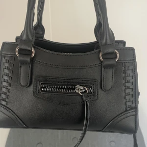 Svart handväska med dragkedja - Snygg svart handväska i skinnimitation från Gina . Väskan har flätade detaljer på sidorna, silverfärgade metalldetaljer och en praktisk dragkedja framtill. Perfekt storlek för det viktigaste och med dubbla handtag för enkel användning. Två fack på insidan, samt ett litet där fram.