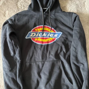Svart Dickies hoodie  - Svart hoodie från Dickies med stor färgglad logga framtill. Klassisk känguruficka och snörning i huvan. Storlek XS, men den är mer som en S. Använd en del och där är en söm i armhålan som gått upp(se sista bild). Därav det billiga priset. Nypris är cirka 700