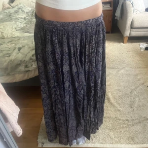 Mönstrad långkjol i marinblått - Säljer en flowy långkjol i marinblått med vitt och vinrött mönster. Kjolen har resår i midjan och är gjord i ett lätt och luftigt material, perfekt för varma dagar. Mönstret är detaljerat och ger en bohemisk vibe. Superskön och snygg till både sneakers och sandaler. Vet ej märke.