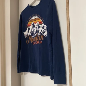 Mörk Blå långärmad tröja H&M Mountain Explorer - Säljer en mörkblå långärmad tröja från H&M med coolt tryck av berg och texten 'Mountain Explorer' i orange och vitt. Rund halsringning och mjukt bomullsmaterial, perfekt för dig som gillar äventyr och snygga prints. Säljer för ett väldigt lågt pris samt hojta till er ifall ni har övriga frågor! 