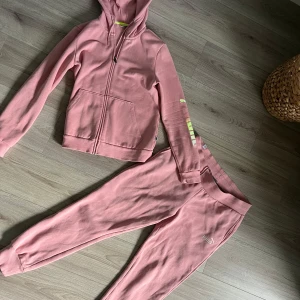 Rosa mjukis set från Puma med tryck - Säljer en rosa mjukis set från Puma med dragkedja, huva och fickor framtill. Ärmen har ett stort Puma-tryck i vitt och limegrönt. Tröjan är i mjuk bomull och har långa ärmar. Perfekt för chill eller träning.