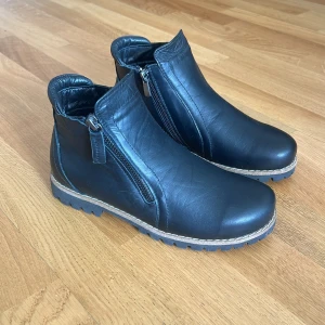 Svarta boots i skinn med dragkedja - Märke: Charlotte och EJ använda!!                            Snygga svarta boots i skinn med grov sula och dragkedja på sidan. Skorna har en rund tå och kontrastsöm längs sulan för en cool look. Perfekta för höst och vinter när du vill ha både stil och komfort. 
