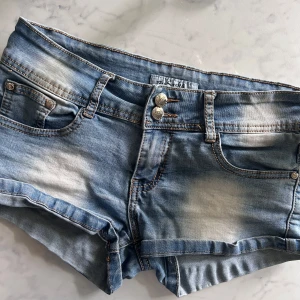 Lågmidjade jeansshorts By Sasha - Snygga ljusblå jeansshorts från By Sasha med låg midja och slitna detaljer. Klassisk femficksmodell med dubbla silverknappar framtill och kontrastsömmar. Perfekta för varma sommardagar och en avslappnad look. S/xs