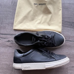 Axel Arigato skor - Stilrena svarta sneakers från Axel Arigato med vit sula och diskret guldfärgad logga på sidan. Skorna har snörning, rund tå och är tillverkade i skinn med detaljer i mocka vid hälen. Perfekta för dig som gillar minimalistisk och modern stil. Skick 7/10. Skicka pm vid minsta fundering