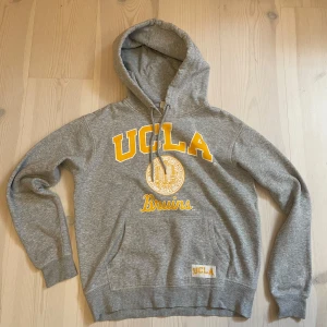 Hoodie | H&M (UCLA). - Grå hoodie från H&M med stor gul UCLA-logga och Bruins-tryck framtill. Klassisk känguruficka, snörning i huvan och mjukt material. Perfekt för dig som gillar college-vibe och sportig stil.