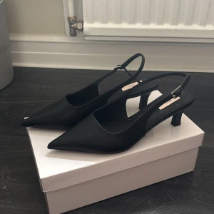 Svarta låga klackskor med spetsig tå från Nelly - Stilrena svarta pumps från Nelly med spetsig tå och låg klack. Skorna har en smal rem med spänne runt hälen och är tillverkade i syntetmaterial. Perfekta för dig som gillar en klassisk och elegant look.