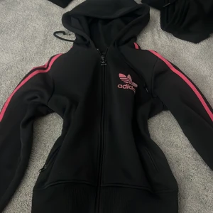 Svart Adidas flis med rosa detaljer - Svart Adidas flis med huva och dragkedja framtill. Rosa ränder längs ärmarna och rosa Adidas-logga på bröstet. Flis har ribbade muddar och fickor med dragkedja. Perfekt för en sportig och avslappnad stil.