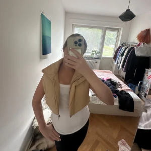 Beige croppad dunväst med dragkedja - Säljer en beige croppad dunväst med hög krage och dragkedja framtill. Västen har en enkel och clean look, perfekt att slänga över en hoodie eller t-shirt. Den är lätt vadderad och har en snygg, boxig passform.