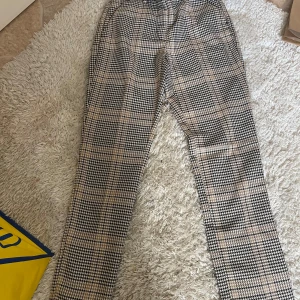 Rutigta kostymbyxor i beige och svart - Snygga kostymbyxor med rutigt mönster i beige och svart. Byxorna har rak passform och klassisk midja, perfekta för en stilren look. Materialet känns mjukt och följsamt, och mönstret ger en trendig vibe.