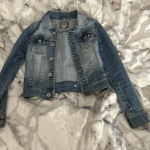 Blå jeansjacka från Detroit - Snygg blå jeansjacka från Detroit med klassisk krage, knappar framtill och två bröstfickor med lock. Jackan har långa ärmar och coola sömdetaljer både fram och bak. Perfekt för dig som vill ha en tidlös och trendig look.