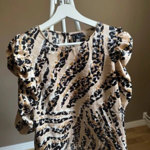 Leopardmönstrad blus från Vero Moda - Snygg blus från Vero Moda med leopardmönster i beige, svart och bruna toner. Modellen har puffiga axlar och trekvartsärm, samt lätt ribbad struktur i tyget. Perfekt för dig som vill sticka ut med ett djurmönstrat plagg.