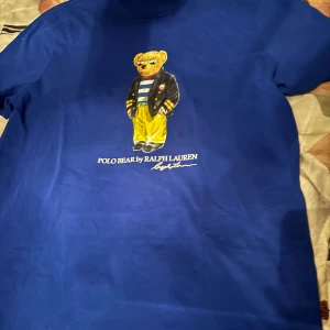 Blå Polo Bear t-shirt från Ralph Lauren - Blå t-shirt från Ralph Lauren med ikoniska Polo Bear-trycket på bröstet. Slim fit-modell i mjuk bomull, kortärmad och med coolt tryck där björnen har randig tröja och solglasögon. Perfekt för dig som vill sticka ut med en klassisk streetstyle. Priset kan diskuteras