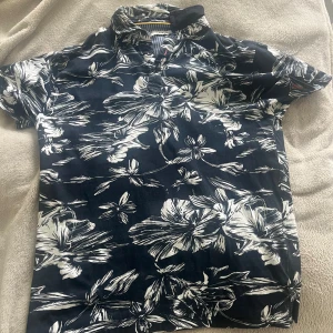 Blommig mörkblå henley från S.C.W - Snygg mörkblå henley-skjorta från S.C.W med stora vita blommor. Skjortan har korta ärmar, knappar vid halsen och en klassisk krage. Perfekt för dig som vill sticka ut med ett coolt mönster och bekväm passform.