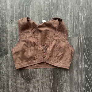 Brun ärmlös linnetopp med knappar - Snygg brun linnetopp med v-ringning och dekorativa spetsdetaljer längs kanterna. Toppen är ärmlös och har fyra knappar fram. Perfekt croppad passform. Materialet är lätt och andas, vilket gör den skön att bära under varma dagar.