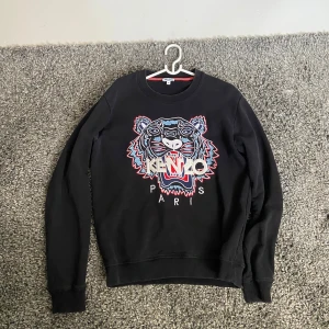 Svart Kenzo sweatshirt med tiger - Svart sweatshirt från Kenzo med ikoniskt broderat tigerhuvud och logga i blått, rött och vitt framtill. Tröjan har rund hals, långa ärmar och är gjord i mjuk bomull. Perfekt för dig som vill sticka ut med en streetstyle-vibe. Sitter som S 