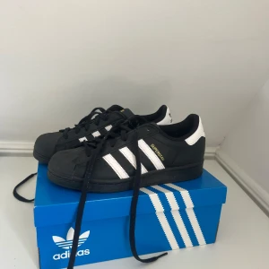 Adidas Superstar svart/vit strl 38 - Klassiska Adidas Superstar sneakers i svart skinn med vita detaljer och de ikoniska tre ränderna på sidorna. Skon har snörning, rund tå och platt sula. Perfekt för dig som gillar en tidlös streetstyle-look.
