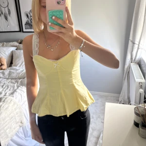 Gul topp med spetsdetaljer - Supersöt gul korsett-topp med vita spetsband och liten rosett framtill. Toppen har figurnära passform upptill och utsvängd nertill. Nästan aldrig använd 