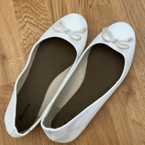 Vita ballerinaskor med rosett från Bata - Snygga vita ballerinaskor från Bata med klassisk rund tå och söt rosettdetalj framtill. Skorna har en platt sula och är tillverkade i ett slätt material som ger en clean look. Perfekta för dig som gillar enkel och stilren design.