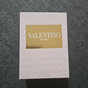 Valentino Donna Eau de Parfum 100ml - Lyxig parfym från Valentino, Donna Eau de Parfum i 100 ml flaska. Flaskan är transparent med ett mönster av små nitar och har en elegant guldfärgad etikett och kork. Doften är feminin och exklusiv, perfekt för dig som gillar stilrena och ikoniska designer.