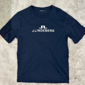 J Lindeberg t shirt - Snygg marinblå t-shirt från J.Lindeberg med vit logga tryckt på bröstet. Klassisk rund halsringning och korta ärmar. Tillverkad i mjuk bomull som känns skön mot huden. Perfekt för dig som gillar att golfa eller bara vill vara skön i vardagen 