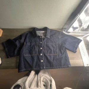 Mörkblå jeansskjorta från Primark - Snygg mörkblå jeansskjorta från Primark med korta ärmar och två stora bröstfickor. Skjortan har klassisk krage, kontrastsömmar och knäppning framtill. Perfekt för en chill och trendig look. Ny pris 200kr 