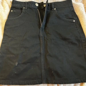 Svart jeanskjol med fickor och dragkedja - Svart kort jeanskjol med klassisk femficksdesign, bälteshällor och dragkedja framtill. Kjolen har raka linjer och bakfickor för en cool och enkel look. Perfekt att matcha med sneakers eller boots.