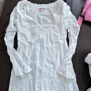 Vit långärmad klänning från Indiska - Superfin vit klänning från Indiska med långärmade ärmar och v-ringning dekorerad med spets. Klänningen har en romantisk känsla med broderade detaljer runt halsen och lätt utsvängda ärmslut. Perfekt för sommarens alla tillfällen!