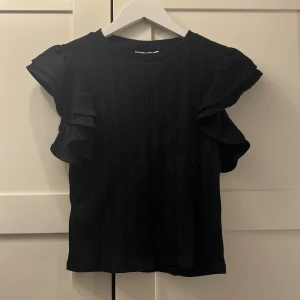 Svart topp med volangärmar - Snygg svart topp med vertikala ribbade ränder och coola volangärmar som ger extra edge. Rund halsringning och korta ärmar med dubbla volanger gör den unik och lätt att styla till olika looks. 