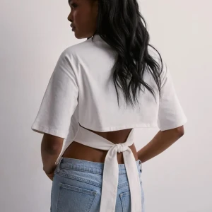 Vit croppad topp med knyt i ryggen - Säljer en vit croppad topp med korta ärmar och snygg knytning i ryggen. Toppen har en enkel och clean look framifrån men en öppen rygg med breda knytband som ger en cool twist. Perfekt till jeans eller kjol för en trendig vibe. Från Nelly i Stl xs, använd men i bra skick💕