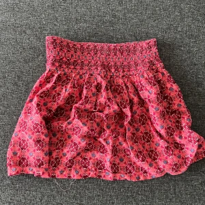 Rosa mönstrad kortkjol med smock - Supersöt kortkjol i rosa med ett färgstarkt mönster och smockad midja. Kjolen har en luftig och flowy passform. Köpt på en marknad i spanien