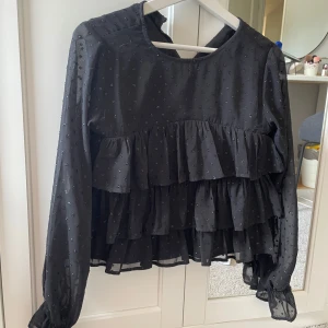 Svart volangblus med prickar - Supersöt svart blus med små prickar och flera lager volanger framtill. Blusen har långärmade, lätt transparenta ärmar med resår vid handlederna och rund halsringning. Perfekt för dig som gillar en romantisk och trendig look. Blusen är knappt andvänd!
