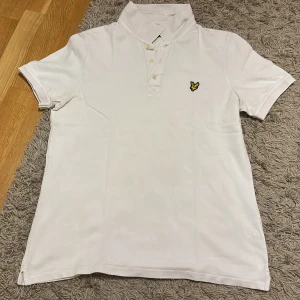Vit pikétröja från Lyle & Scott XL - Klassisk vit pikétröja från Lyle & Scott i storlek XL. Tröjan har korta ärmar, krage och knappar framtill samt den ikoniska gula örnloggan broderad på bröstet. Tillverkad i mjuk bomull som känns skön mot huden. Perfekt för en clean och stilren look.
