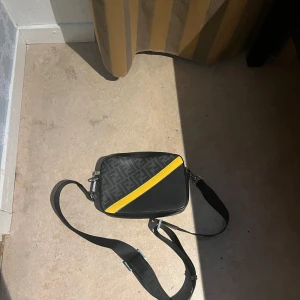 Fendi axelväska med gul detalj - Snygg axelväska från Fendi i svart skinn och textil med klassiskt FF-mönster. Väskan har en gul diagonal detalj på framsidan och justerbar axelrem. Perfekt storlek för det viktigaste och ger en cool vibe till din outfit. Använd 2-3 gånger