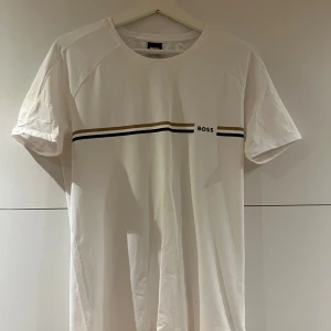 Vit t-shirt från BOSS med ränder - Vit t-shirt från BOSS med stilrena svarta och guldiga ränder över bröstet och diskret logga. Klassisk passform och rund halsringning, perfekt för en clean och snygg look.