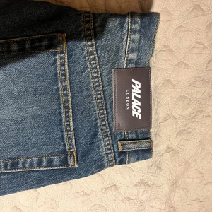 Palace London blå jeans straight fit - Säljer ett par blå jeans från Palace London med klassisk femficksdesign och snyggt slitna detaljer framtill. Jeansen har rak passform och normal midja, tillverkade i robust denim med kontrastsömmar och stor logopatch bak i midjan. Köptes i våras sällan använda