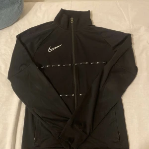 Svart Nike Dri-Fit träningsjacka S - Svart Nike Dri-Fit träningsjacka med hel dragkedja och högt krage. Jackan har vita Nike-loggor som band runt bröstet och ryggen, samt två fickor med dragkedja. Tillverkad i lätt och stretchig polyester, perfekt för sportiga dagar.