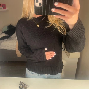 Svart stickad tröja  - En svart stickad tröja med rund hals och breda ribbade muddar vid ärmslut och nederkant. Tröjan har en avslappnad passform och är långärmad, perfekt att matcha med jeans för en enkel och stilren look.