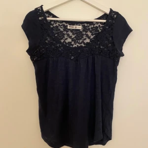Mörkblå blus med spets från Svea - Supersöt mörkblå blus från Svea med vacker spets upptill och korta ärmar. Blusen har rund halsringning och ett romantiskt blommigt spetsmönster över axlar.