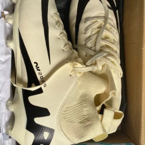 Beige och svarta fotbollsskor Air 2025 - Snygga fotbollsskor i beige med svarta detaljer och coolt Air 2025-tryck på sidan. Skorna har snörning, stickad ovandel och plös med dragflik för enkel påtagning. Perfekta för dig som vill sticka ut på planen.